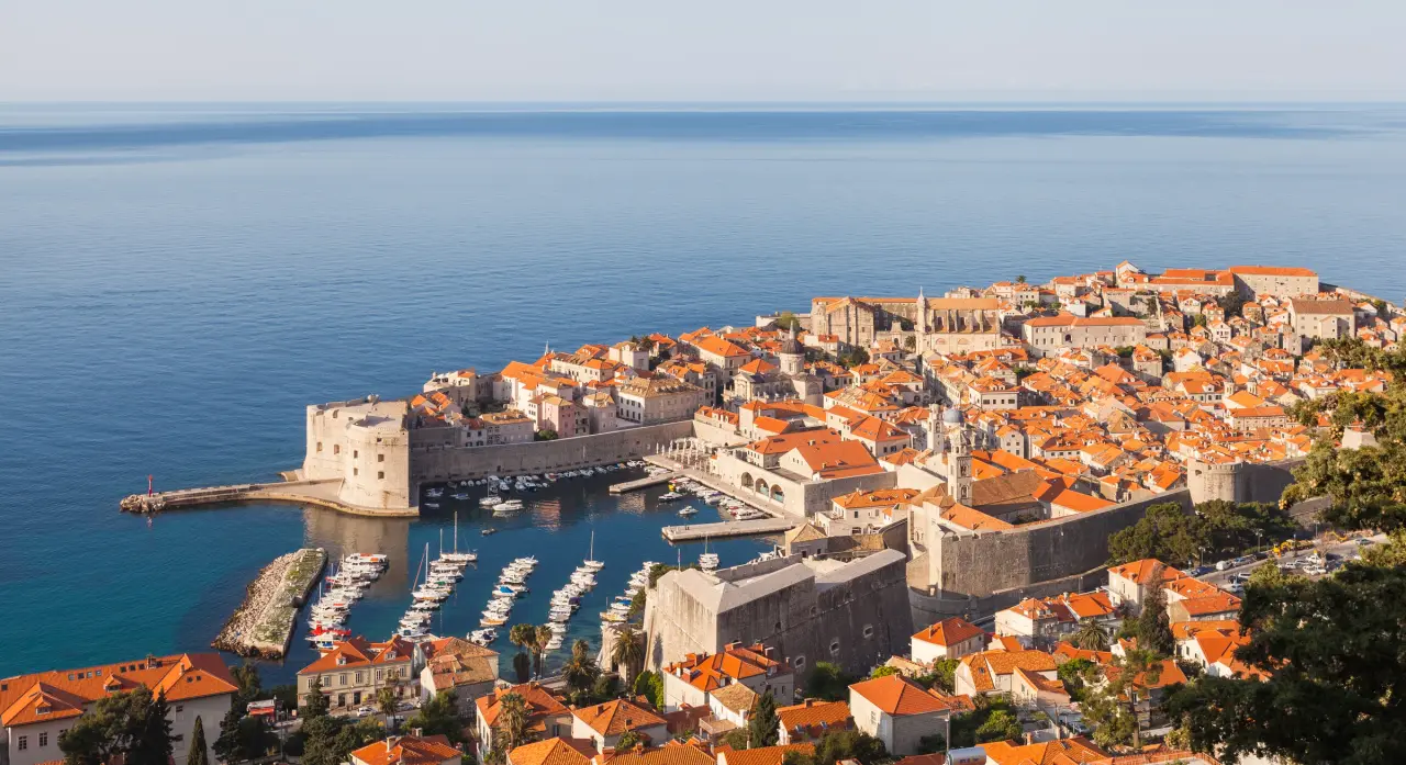 Sonnenbeschienene Mauern und orangefarbene Dächer der Altstadt von Dubrovnik neben der blauen Adria und dem Hafen.