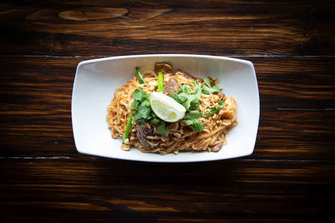A plate of stir-fried noodles is topped with herbs and a wedge of lime.