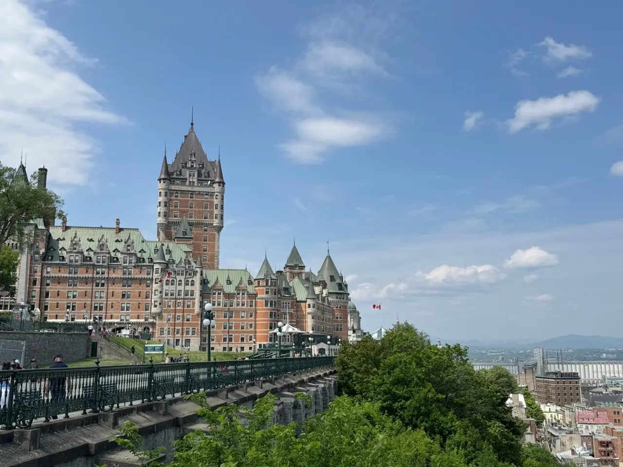 Château Frontenac rises above the public promenade and green treetops in Québec City beneath a clear blue summer sky.