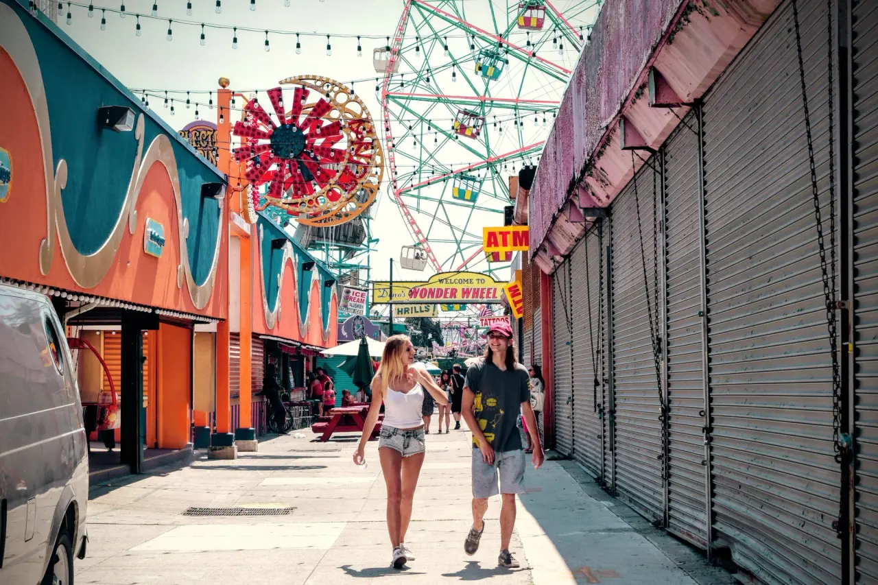 Deux visiteurs passent devant des façades colorées de parc d'attractions et des promenades par une journée ensoleillée.