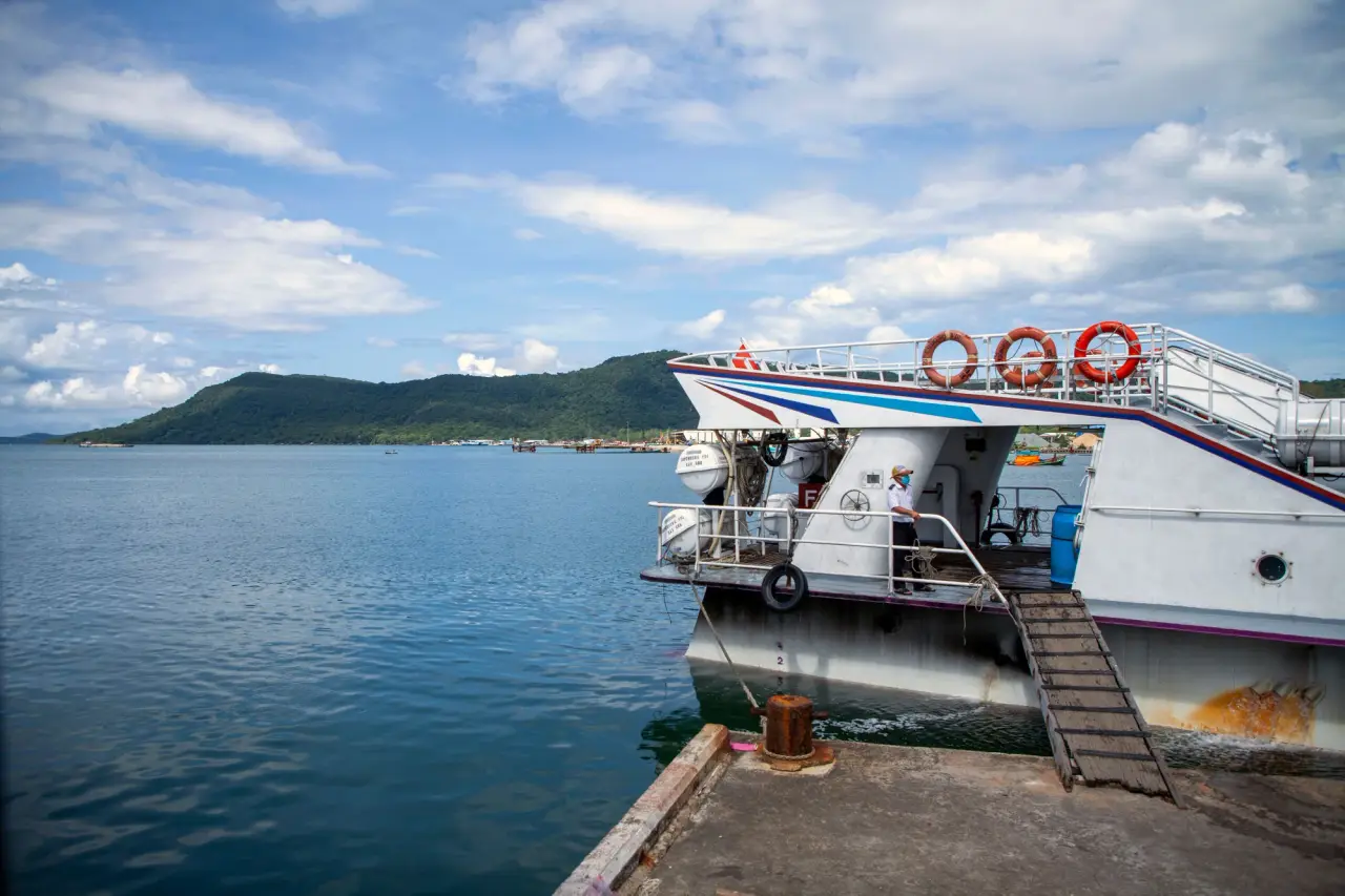 Un ferry al lado de un muelle tiene una pasarela de embarque, con agua azul y una verde colina costera más allá.