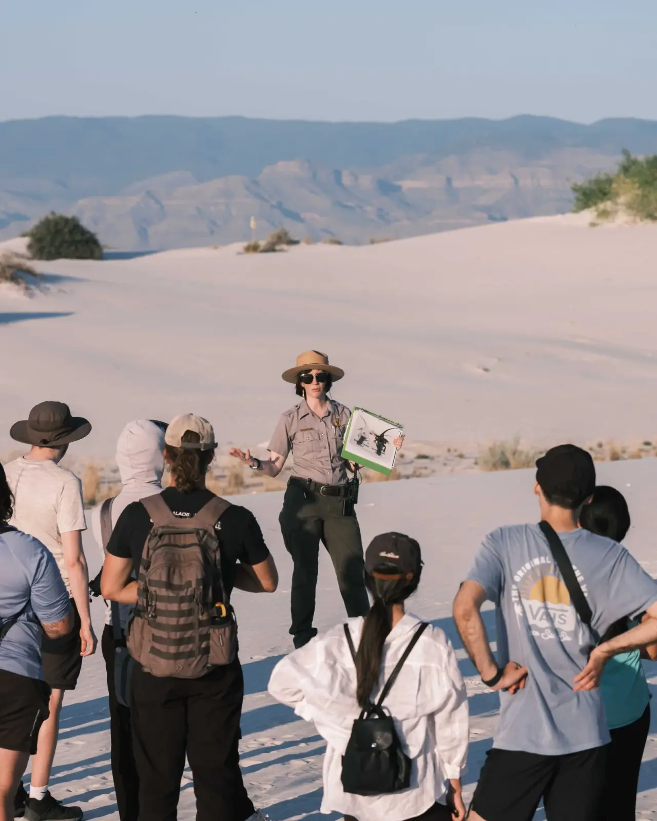 A park ranger holds an illustrated guide while speaking to visitors among white sand dunes.