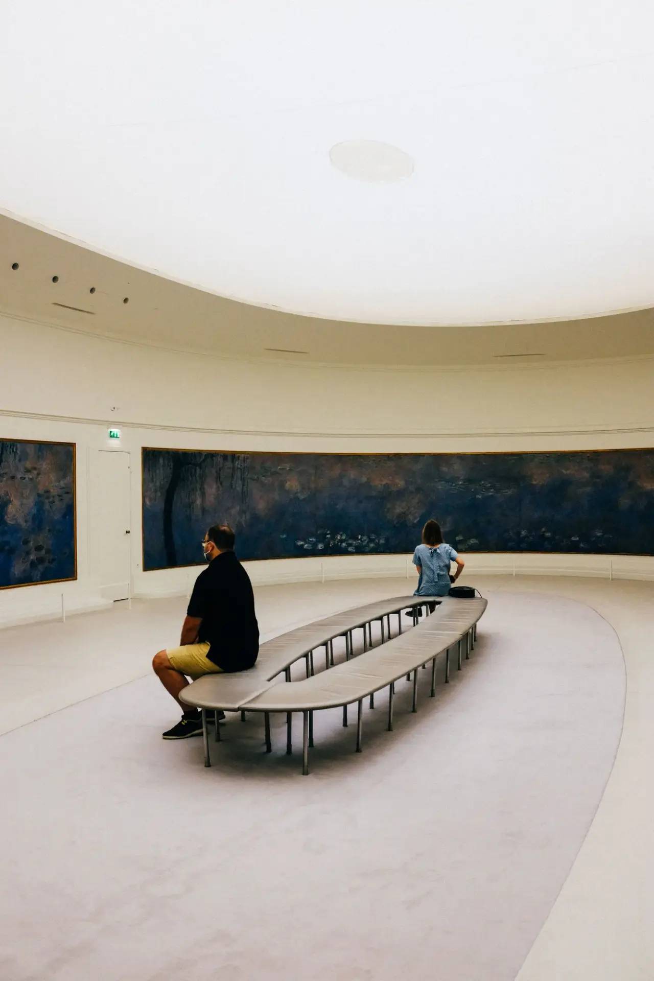 Besucher sitzen auf einer geschwungenen Bank vor einem langen Gemälde in einer hellen, runden Galerie.