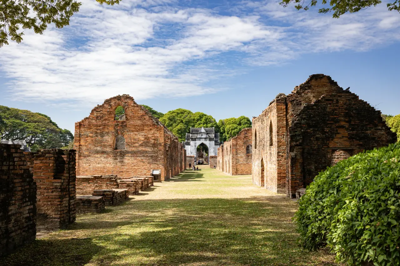 Sunlit brick ruins and a grassy passage lead toward a white arched gate inside King Narai’s Palace in Lopburi.