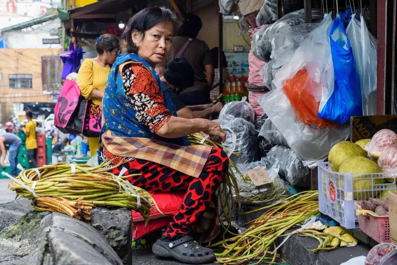 A market seller sits among colourful vegetables and produce bags at an outdoor stall.