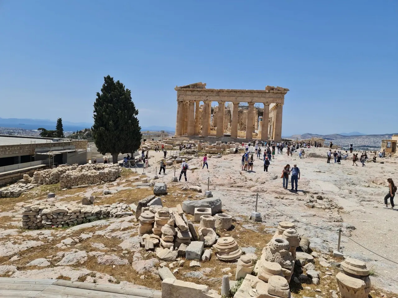 Des visiteurs suivent une allée en pierre claire vers le Parthenon sous un ciel bleu dégagé.