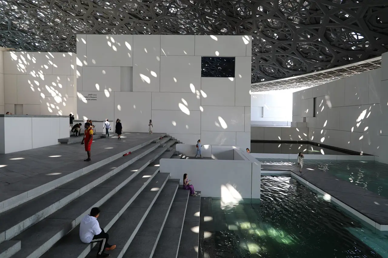 Sunlight filtering through the Louvre Abu Dhabi dome onto white gallery buildings, steps and water.
