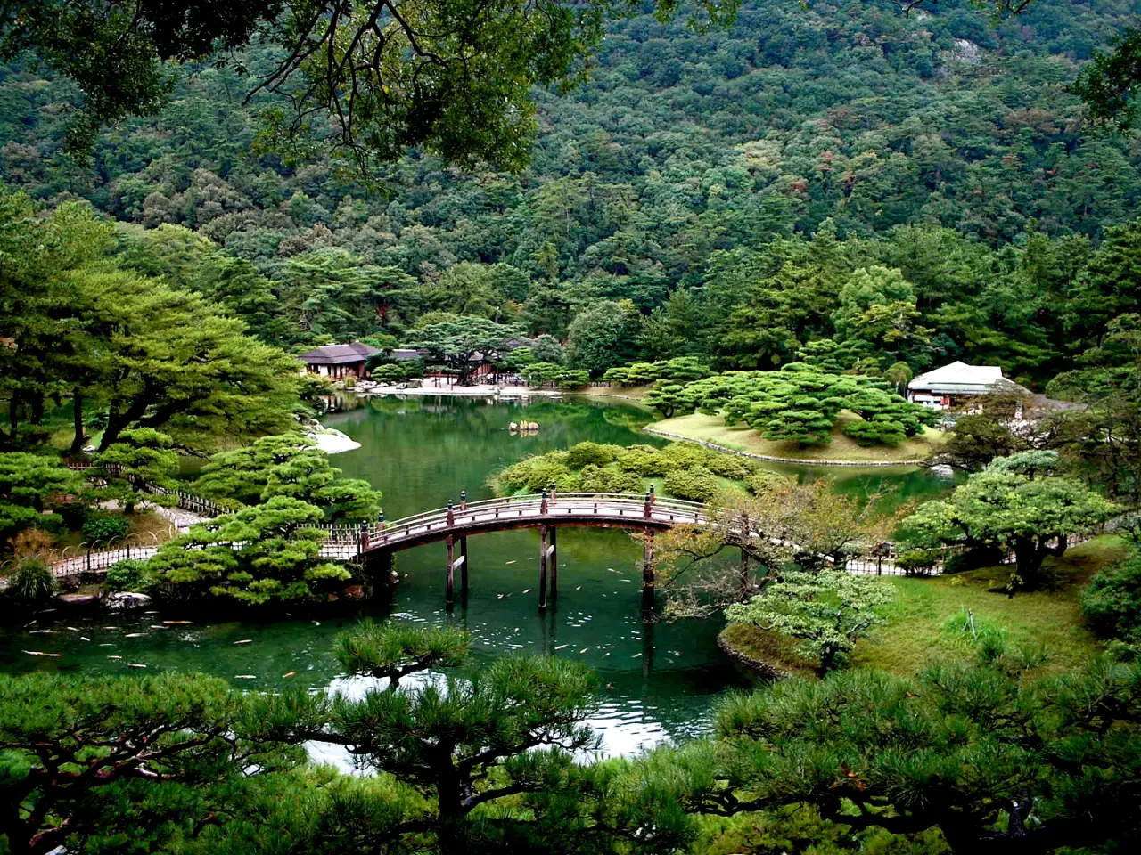 Green pines, wooded hills and reflections around a pond in Ritsurin Garden, with an arched bridge along the landscaped shore.