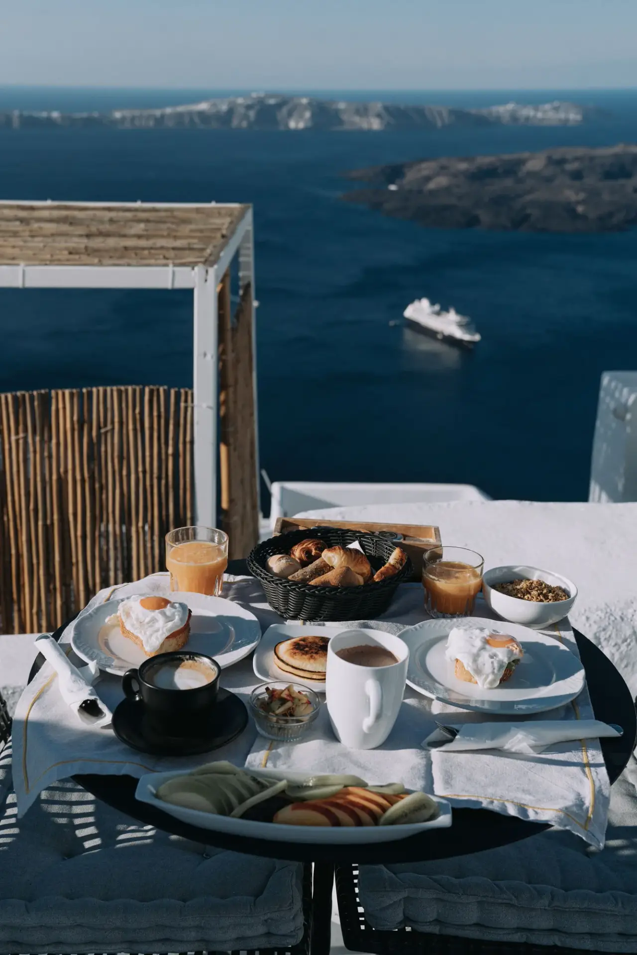A breakfast table with pastries, juice and coffee overlooks a deep blue sea from a hotel balcony.