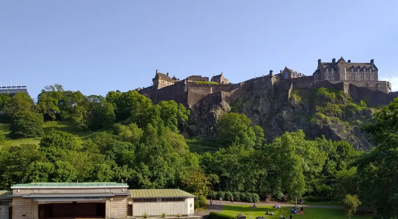 Sunlit main buildings of Edinburgh Castle above green Princes Street Gardens.