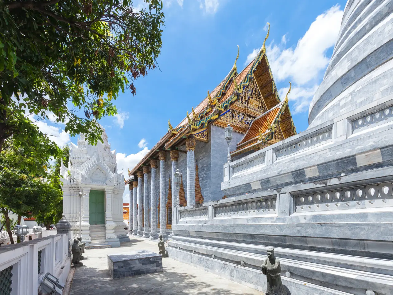 ผนังหินอ่อนรับแสงแดดและรายละเอียดหลังคาสีทองที่วัดราชประดิษฐ์ในกรุงเทพฯ ข้างต้นไม้ใบดก