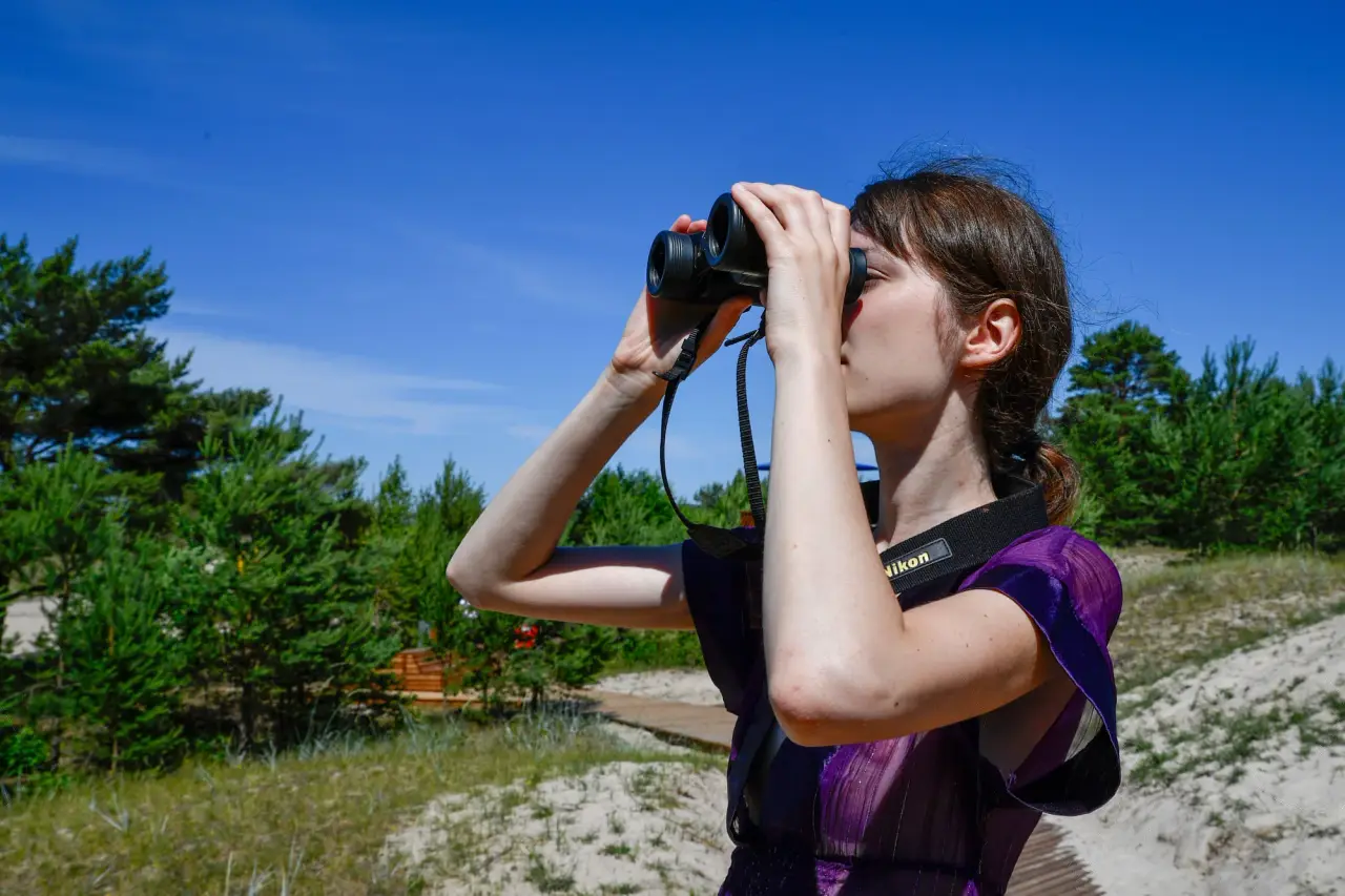 Une femme dans un haut violet regarde à travers des jumelles à côté d'arbres verts bas sous un ciel bleu.