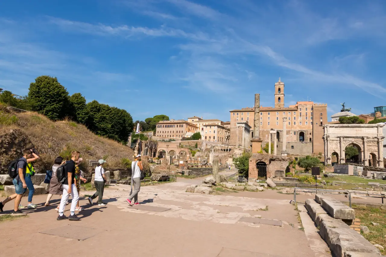 Visitors walk among the Roman Forum ruins in Rome beneath a clear blue sky.