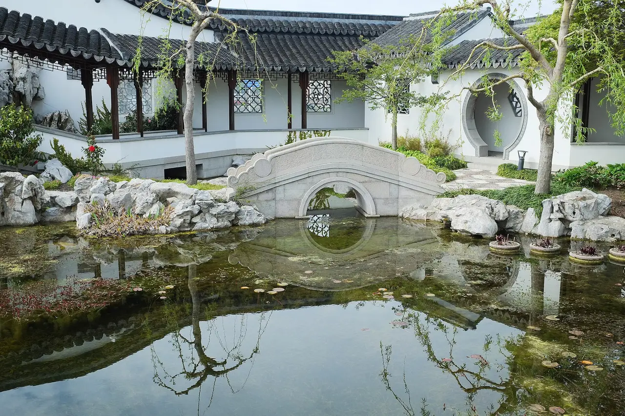 Eine gewölbte Steinbrücke, die sich im Teich des chinesischen Gartens von Dunedin widerspiegelt, mit traditionellen Pflanzungen und Hofmauern