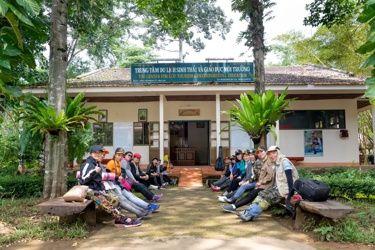 Visitors rest on benches outside an ecotourism information building surrounded by green trees.