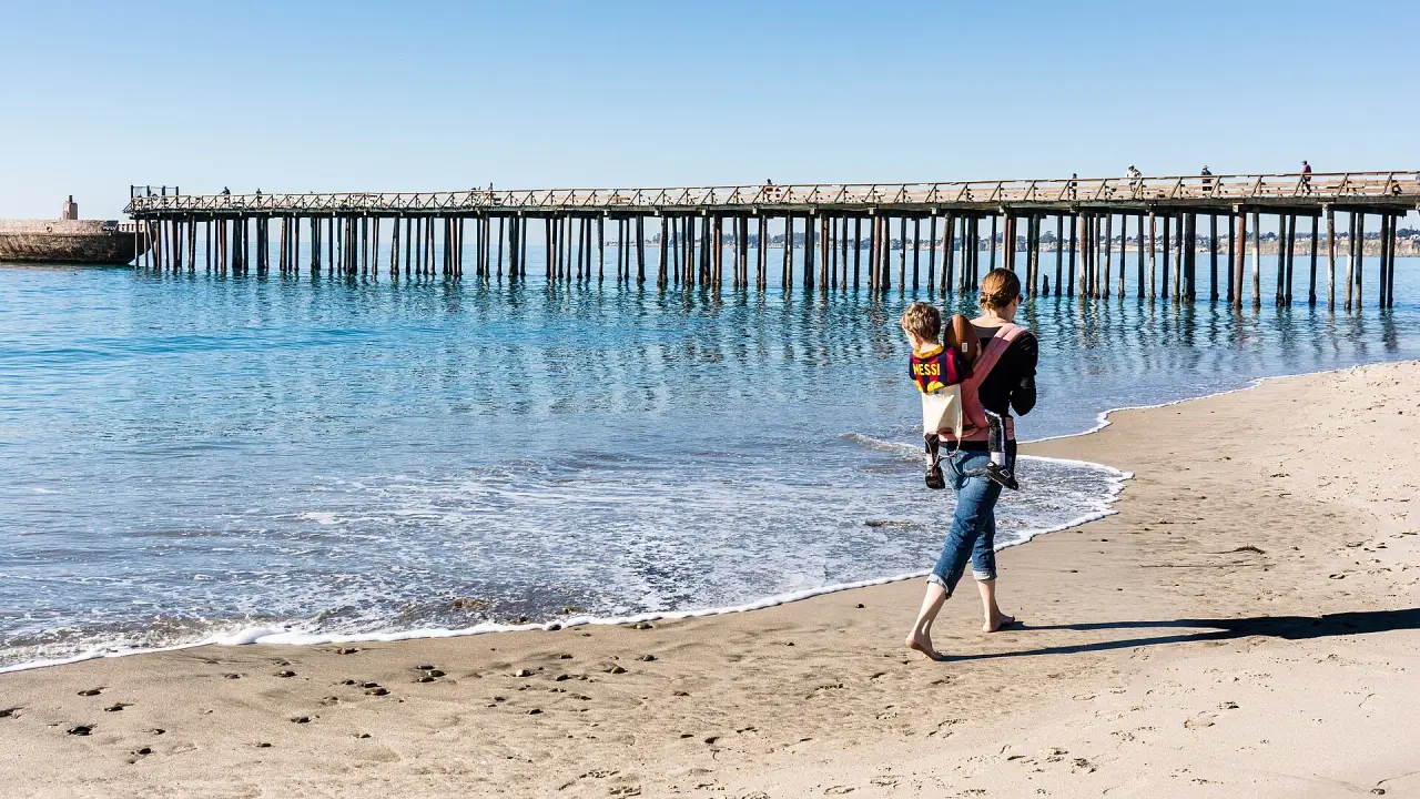 A parent carrying a child in a backpack beside a sunny beach pier