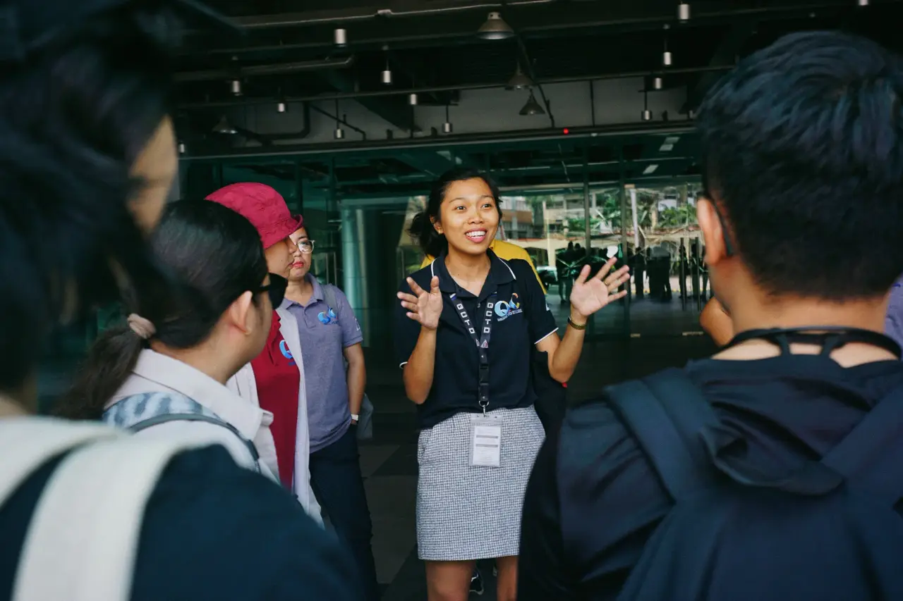 A woman gestures with both hands while briefing a small group outdoors.