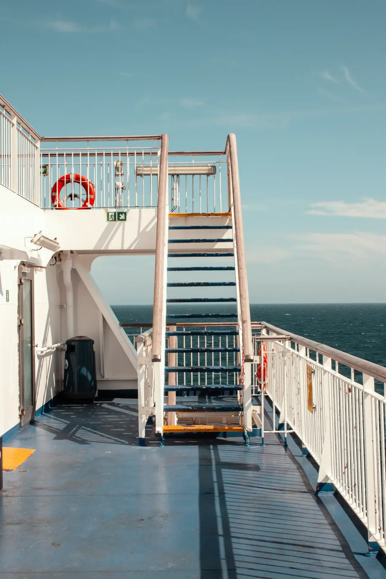 A painted stairway rises from an open ferry deck beside calm sea water.