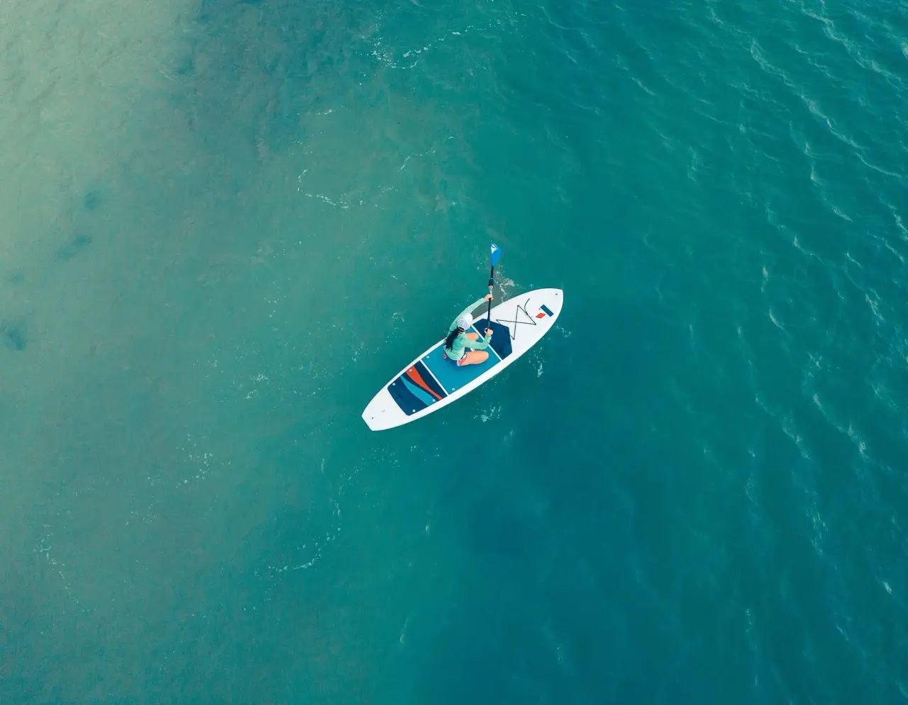 An aerial view shows a paddler on a white paddleboard in clear turquoise water.