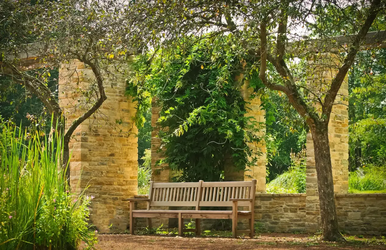 A wooden bench stands between stone garden arches under leafy trees.