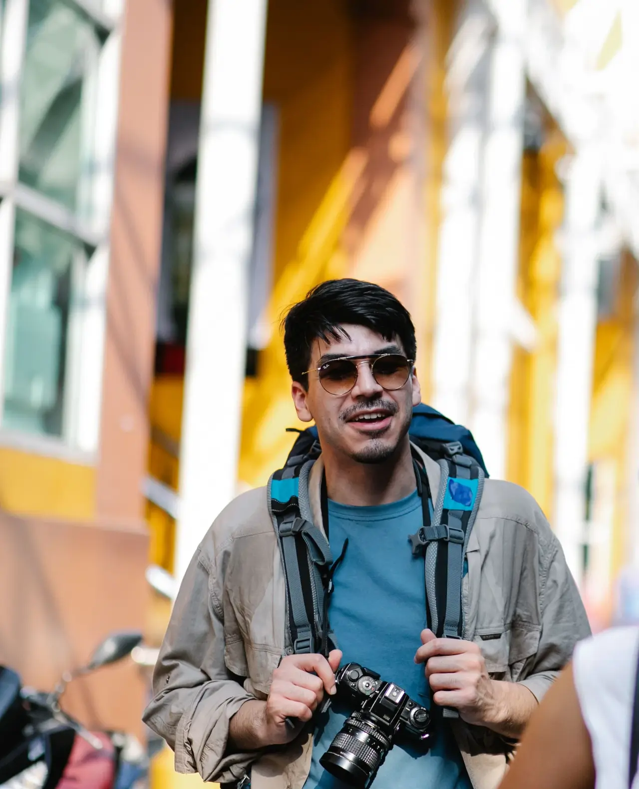 A visitor wearing sunglasses and a backpack walks through a colourful street market.