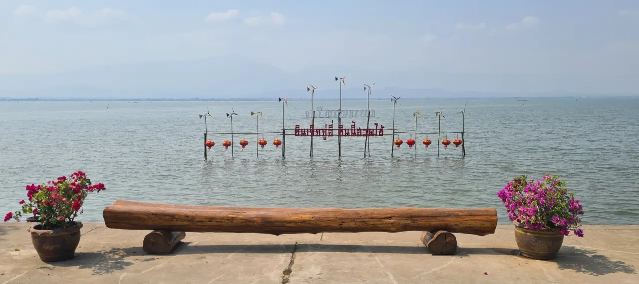 A wooden bench and flowering pots overlook Kwan Phayao, with a Thai lakeside sign standing in the water.