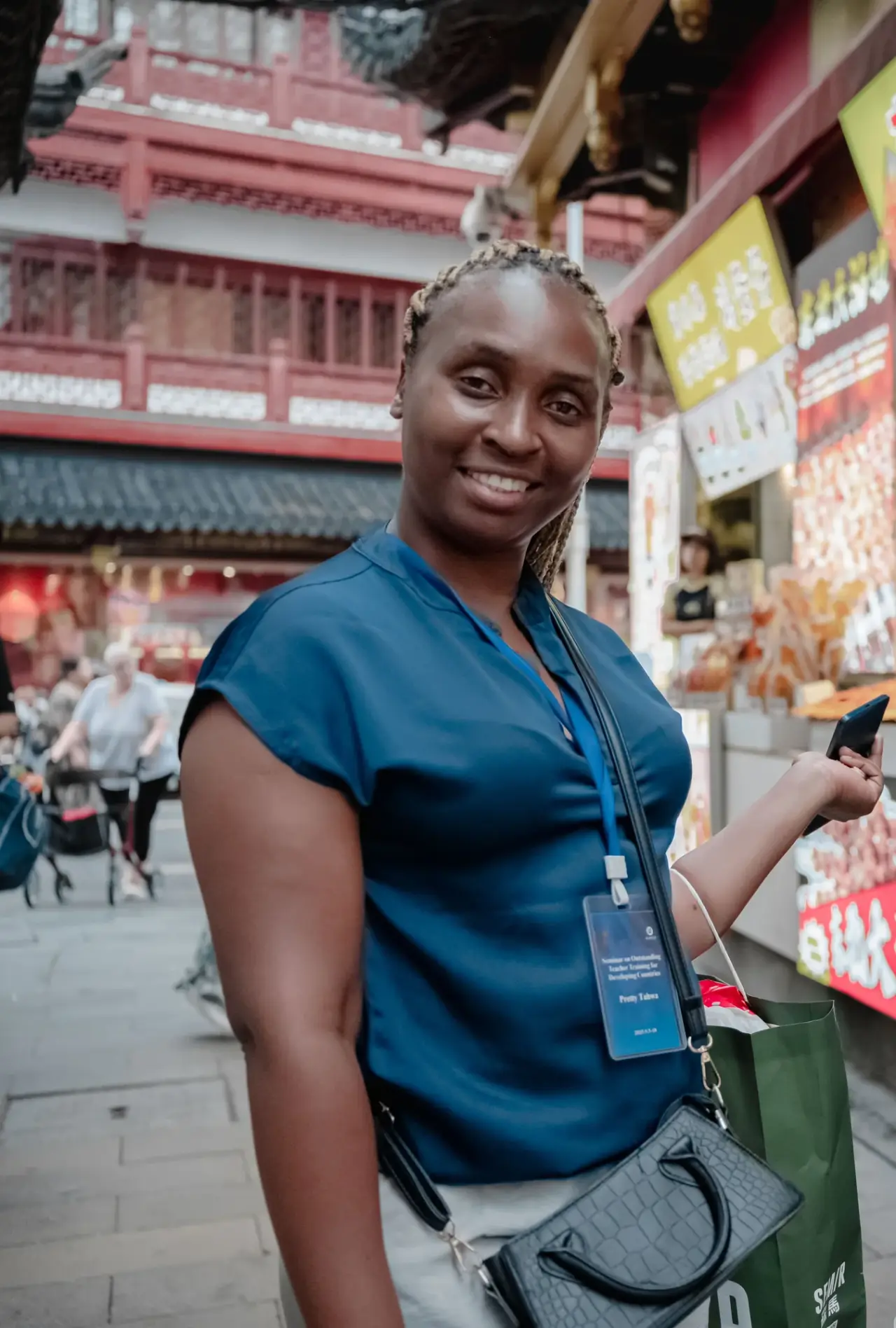 A smiling visitor in a blue top stands among colourful market signs and a red building.