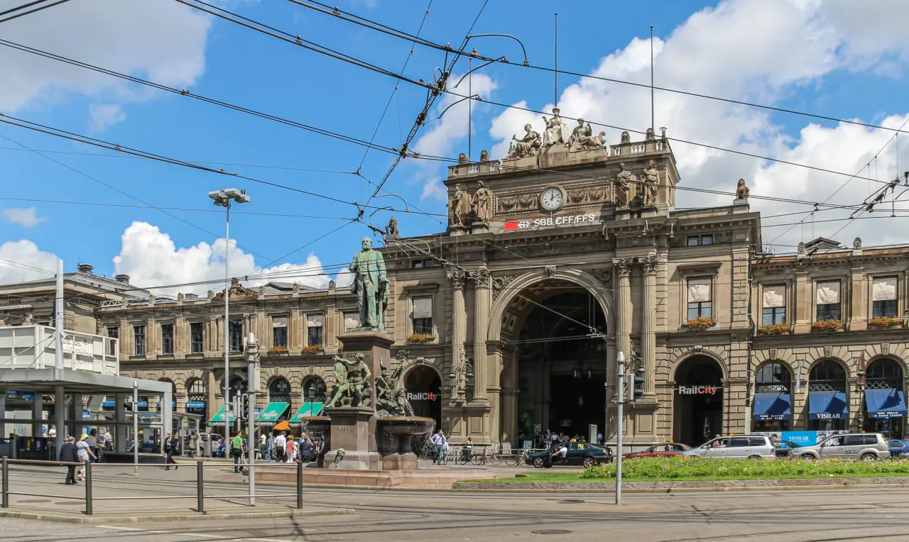 The main entrance of Zürich Hauptbahnhof under blue sky, with tram wires and travellers in front.