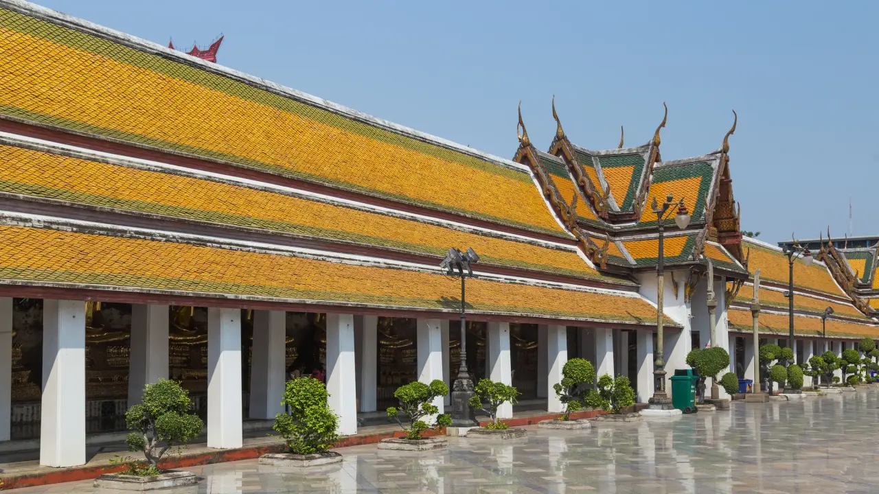 Golden-tiled cloister roofs and an ornate entrance at Wat Suthat in Bangkok beside a sunlit courtyard.