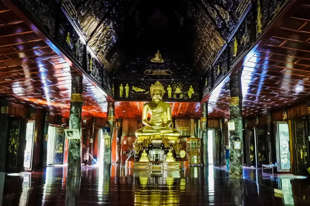 A golden seated Buddha and altar between timber columns on the reflective floor inside Wat Phuttha Nimit in Kalasin.