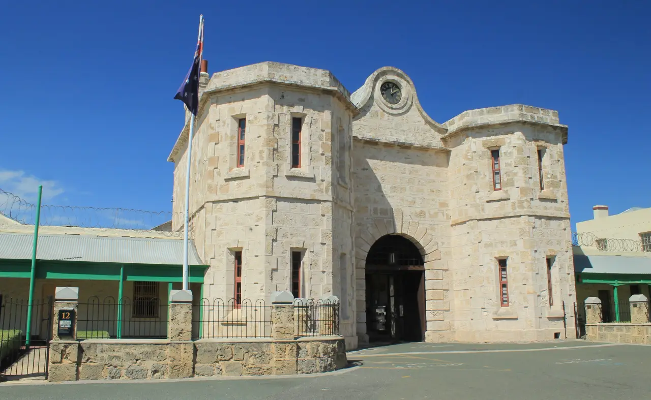 Sonnenbeschienenes Torhaus aus Kalkstein des Fremantle Prison mit zwei Türmen, einem Torbogen und einer Uhr vor blauem Himmel.