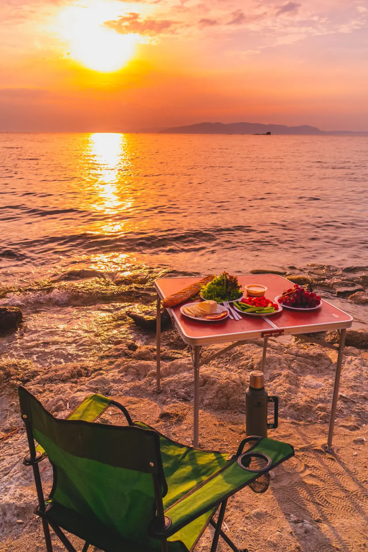 A small picnic table is set on a beach beside sunset-lit water.