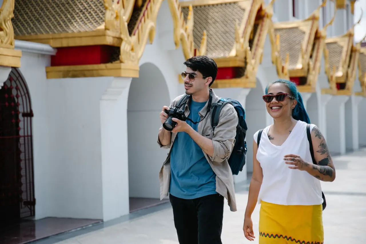Dos viajeros con cámaras caminan junto a un edificio ornamentado de un templo tailandés.